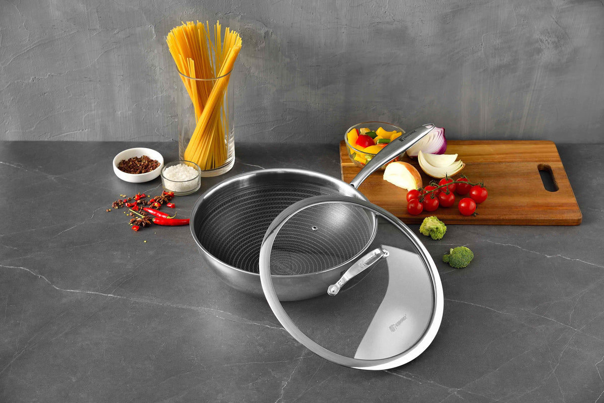 A stainless steel sauté pan with a matching glass lid is displayed on a dark gray countertop, surrounded by cooking ingredients including spaghetti in a glass, a cutting board with vegetables, and small bowls of spices.slider_item_AaQpUp